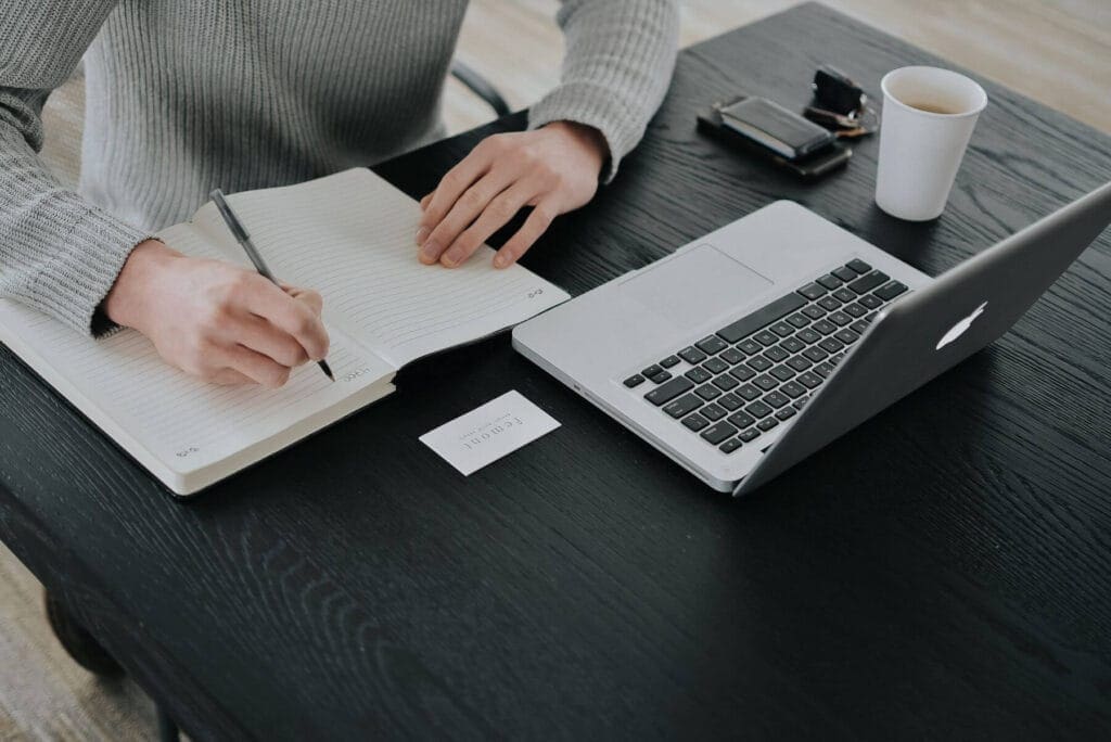 person writing in notebook with laptop at desk